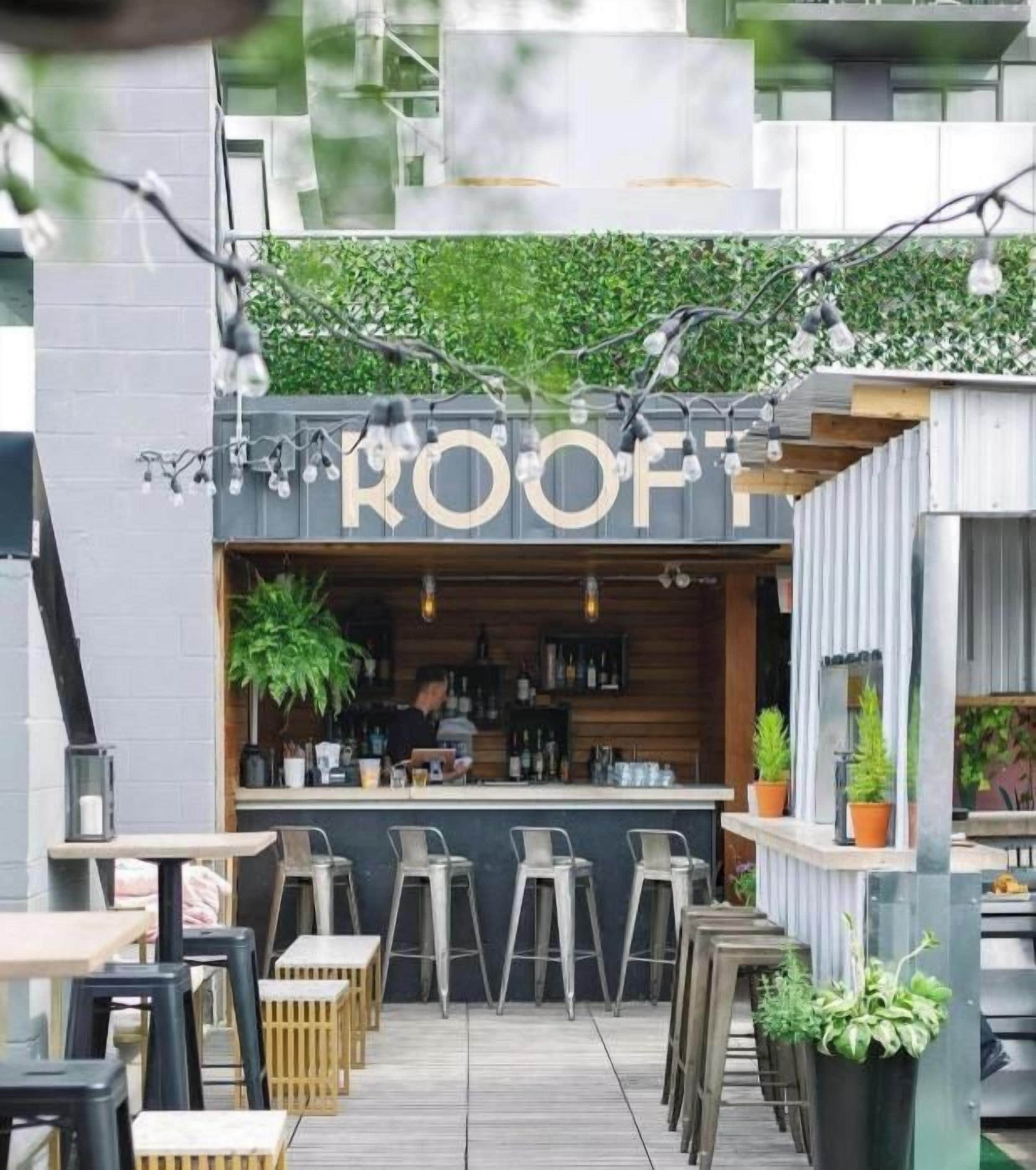 An outdoor bar area with a sign reading 'ROOF' and a bartender behind the counter. There are multiple seating areas with stools and tables.