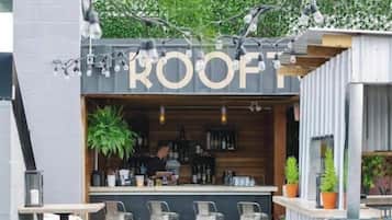 An outdoor bar area with a sign reading 'ROOF' and a bartender behind the counter. There are multiple seating areas with stools and tables.
