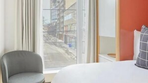 Habitación de hotel con una cama grande, una silla, una ventana con cortinas y vistas a edificios.