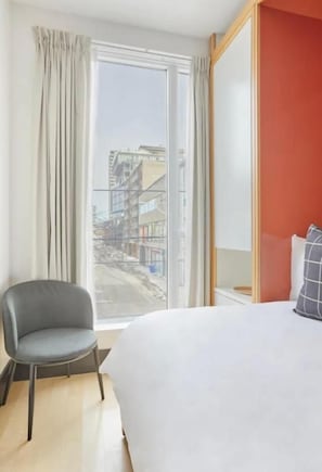 Quarto de hotel com uma cama grande, uma cadeira, uma janela com cortinas e vista para prédios.