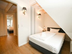 A bedroom with a sloped ceiling, a bed with white linens, two wall-mounted lamps, and a visible door leading to another room. - Bonde Hus Henny (Büsum)