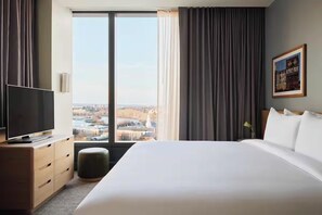 A hotel room with a large bed, a television on a wooden dresser, and a view of a cityscape through a large window.
