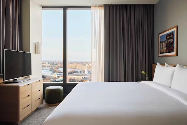 A hotel room with a large bed, a television on a wooden dresser, and a view of a cityscape through a large window.