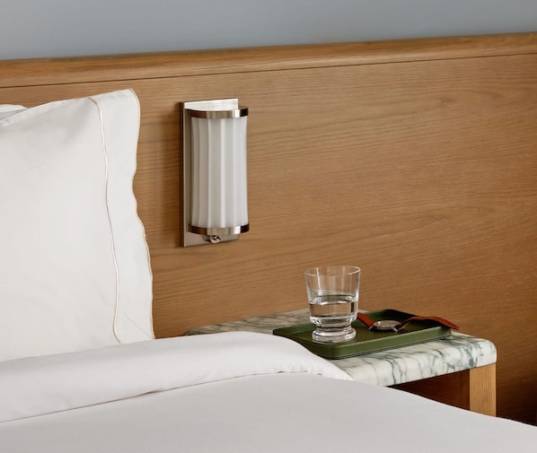 Ein Hotelzimmer mit einem Holzkopfteil, einer weißen Bettdecke, einem Marmornachttisch, einem Glas Wasser und einer grünen Ledertasche.