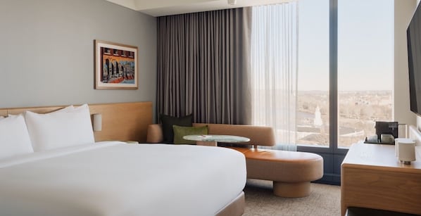 A hotel room with a large bed, a small round table, a sofa, and a desk with a lamp. There is a window with a view of the city. - King Room | The Atlas Hotel | 0.4 Miles to Harvard Stadium (Boston)
