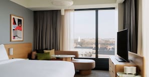 Ein Hotelzimmer mit einem großen Bett, einem Sofa, einem kleinen Tisch, einem Fernseher und einem Fenster mit Blick auf die Stadt.