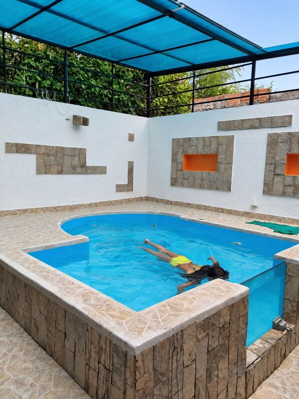 A swimming pool with a person swimming, surrounded by stone walls and a blue canopy.