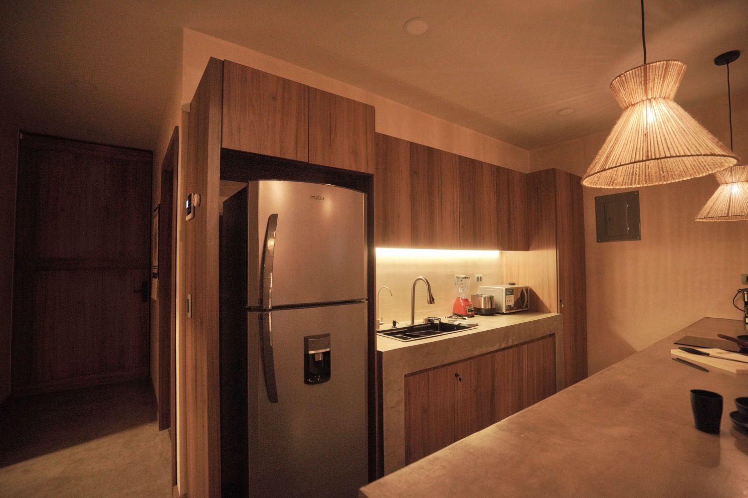 Cozinha moderna com armários de madeira, geladeira de aço inoxidável, pia embutida e luminária pendente.