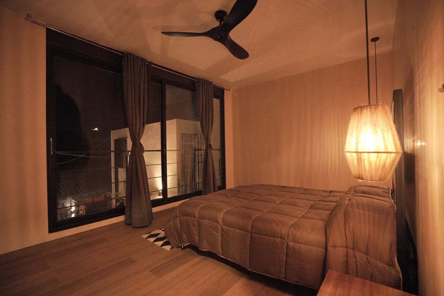 Quarto com cama, ventoinha no teto, uma janela grande com cortinas e abajur.