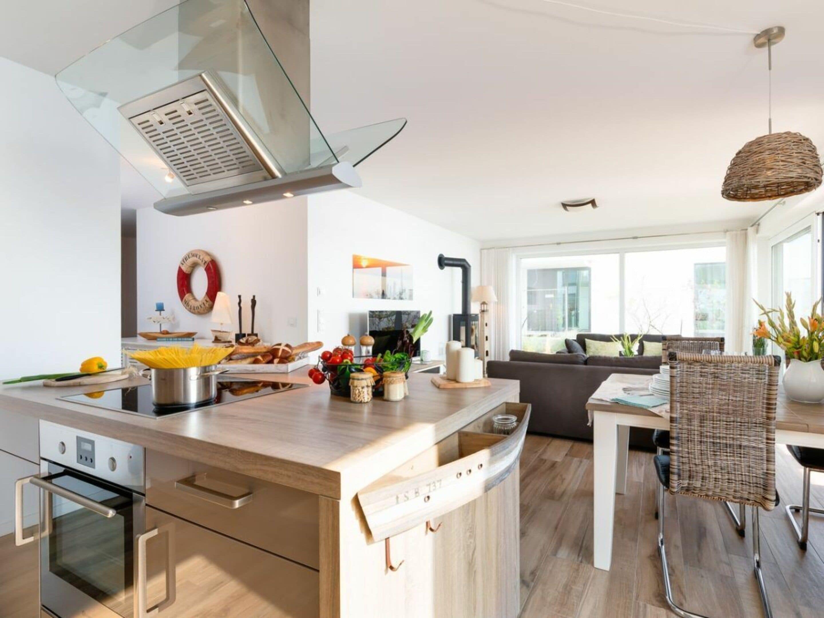 Una cocina moderna con una isla central, horno empotrado y un comedor con una silla de mimbre y un florero.