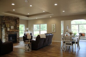A spacious living room with a fireplace, leather sofas, a dining area with a table and chairs, and large windows offering a view of greenery.
