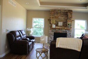 A living room with a stone fireplace, leather sofas, and a view of trees through the windows. - Enjoyable 5-bedroom house with AC in welcoming Wellman (Wellman)