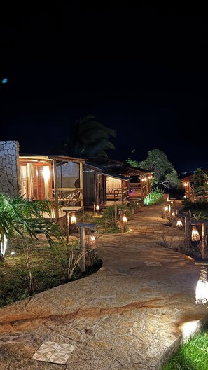 A pathway leading to a rustic building with a stone facade, wooden deck, and ambient lighting.