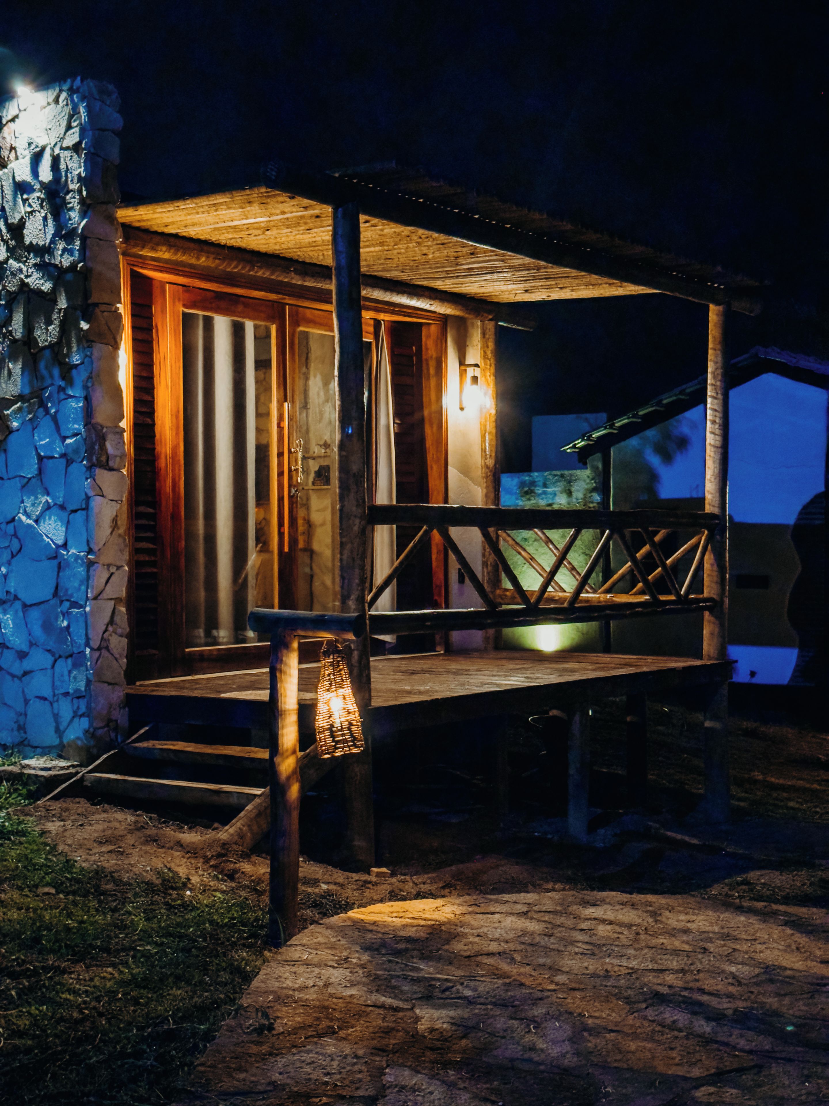 A rustic wooden cabin with a porch, stone wall, and a small light fixture.