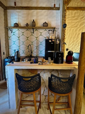 A kitchen with a stone counter, two wicker barstools, a coffee machine, and a refrigerator.