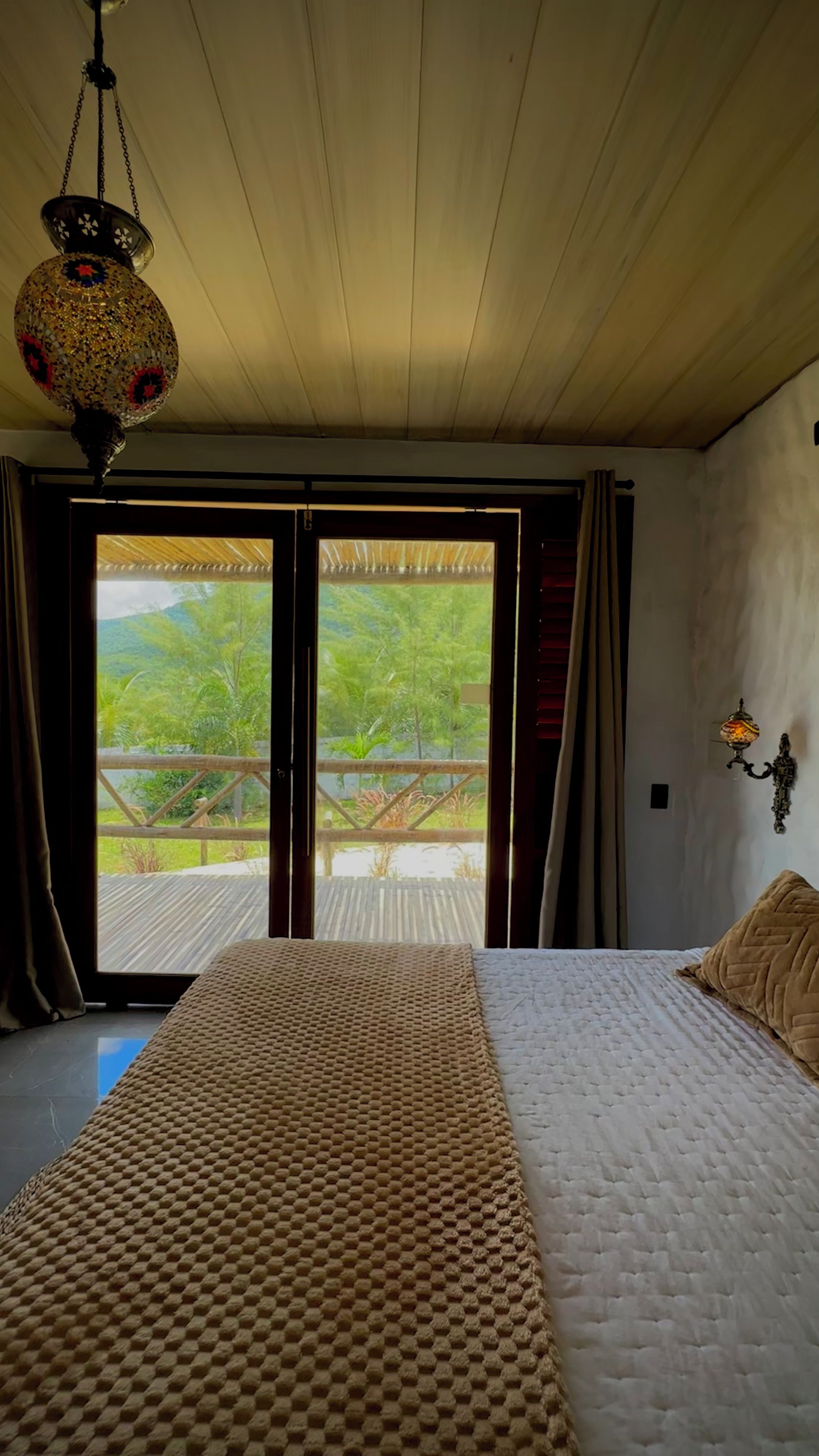 Quarto com cama, luminária pendente e vista para um deck de madeira e vegetação.