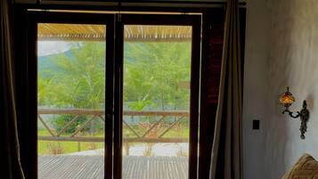 Quarto com cama, luminária pendente e vista para um deck de madeira e vegetação.