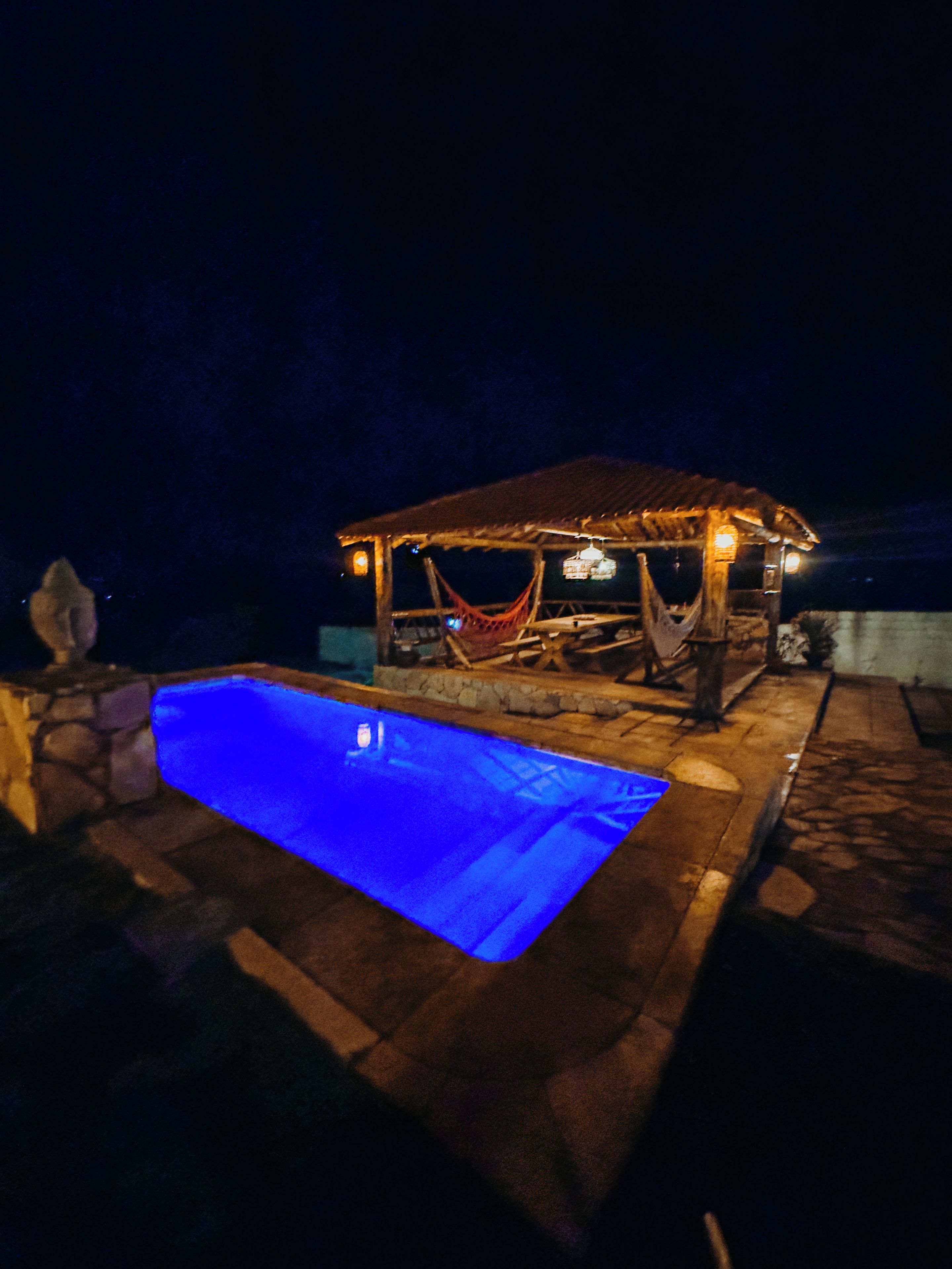 Un área de piscina exterior con piscina iluminada por la noche, una hamaca y una pequeña estructura cubierta.