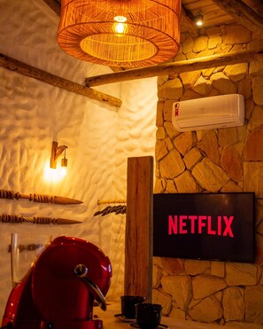 A room with a stone wall, an air conditioning unit, a mounted television displaying 'NETFLIX', and a red appliance on the counter.