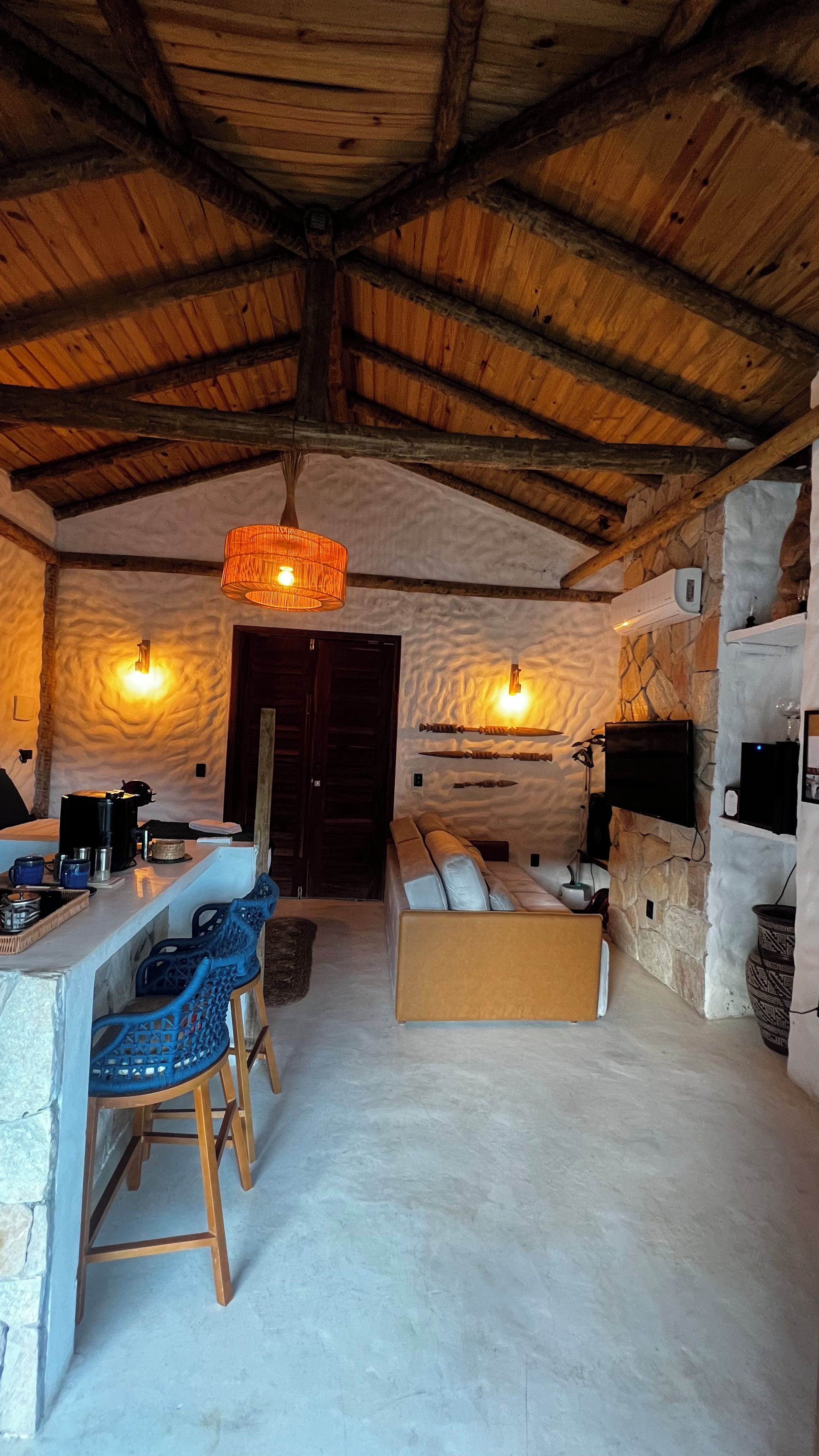 Un interior rústico con pared de piedra, techo de madera, un sofá, una zona de bar con taburetes y un televisor montado en la pared.