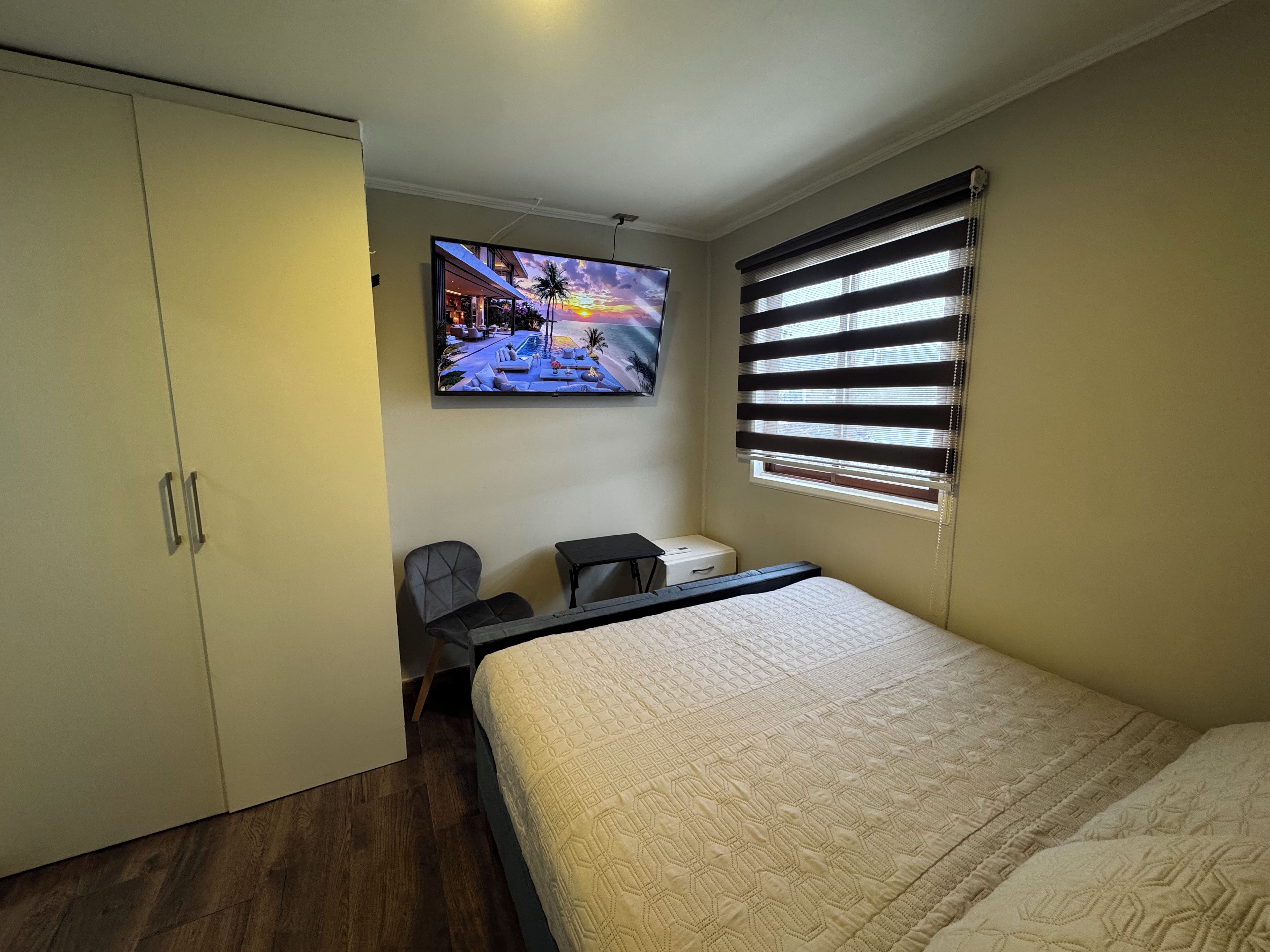Quarto com cama, escrivaninha, cadeira, guarda-roupa e televisão exibindo uma cena de pôr do sol.