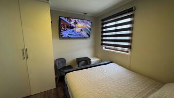 Quarto com cama, escrivaninha, cadeira, guarda-roupa e televisão exibindo uma cena de pôr do sol.
