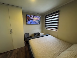A bedroom with a bed, a desk with a chair, a wardrobe, and a television displaying a sunset scene.