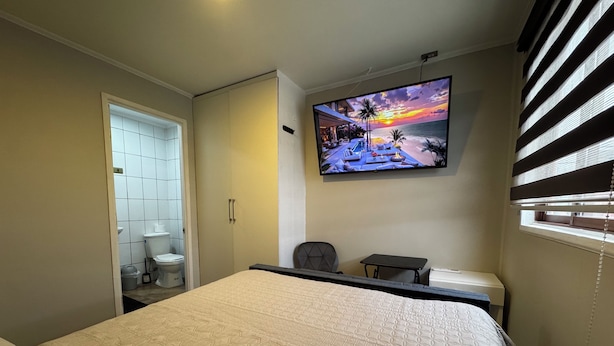 Une chambre d’hôtel avec un lit, un téléviseur fixé au mur, une petite table et une fenêtre avec des stores.