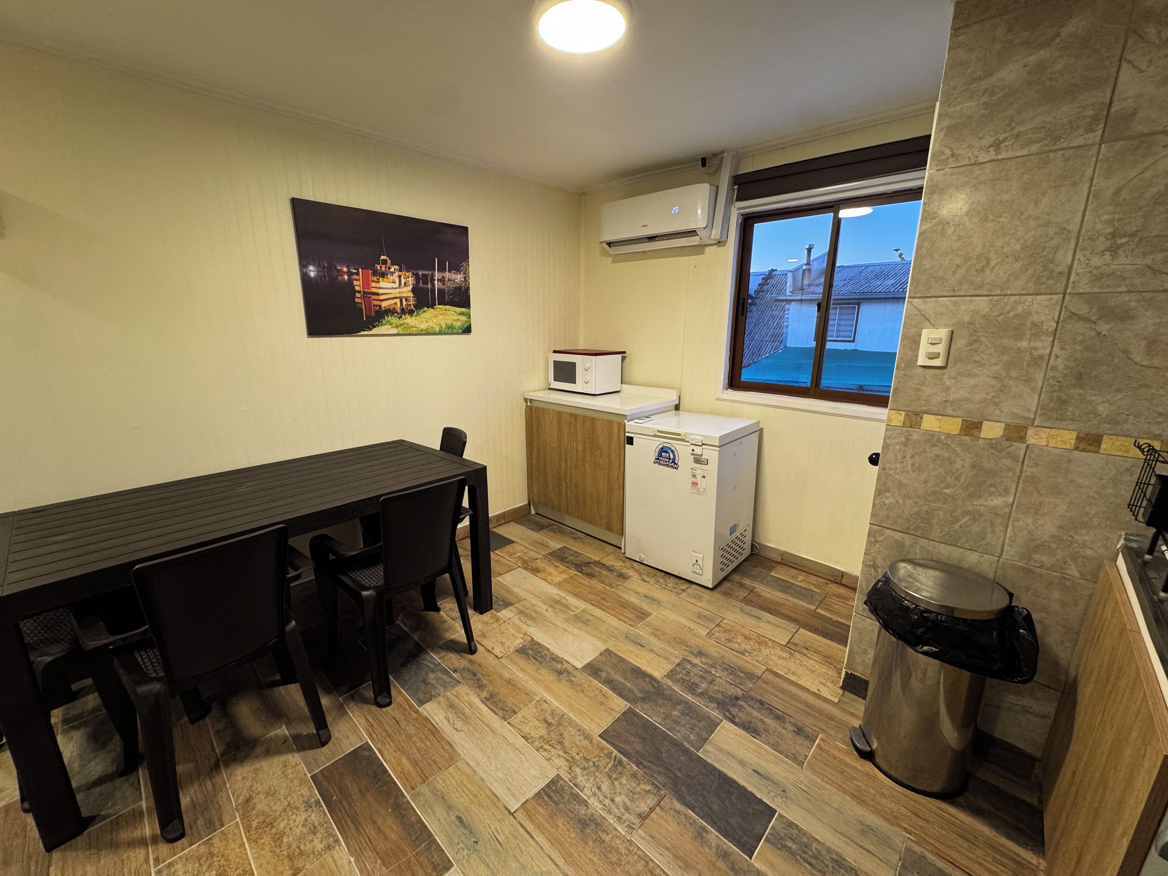 Cocina compacta con mesa, sillas, microondas, refrigerador pequeño, ventana con vistas y un basurero.