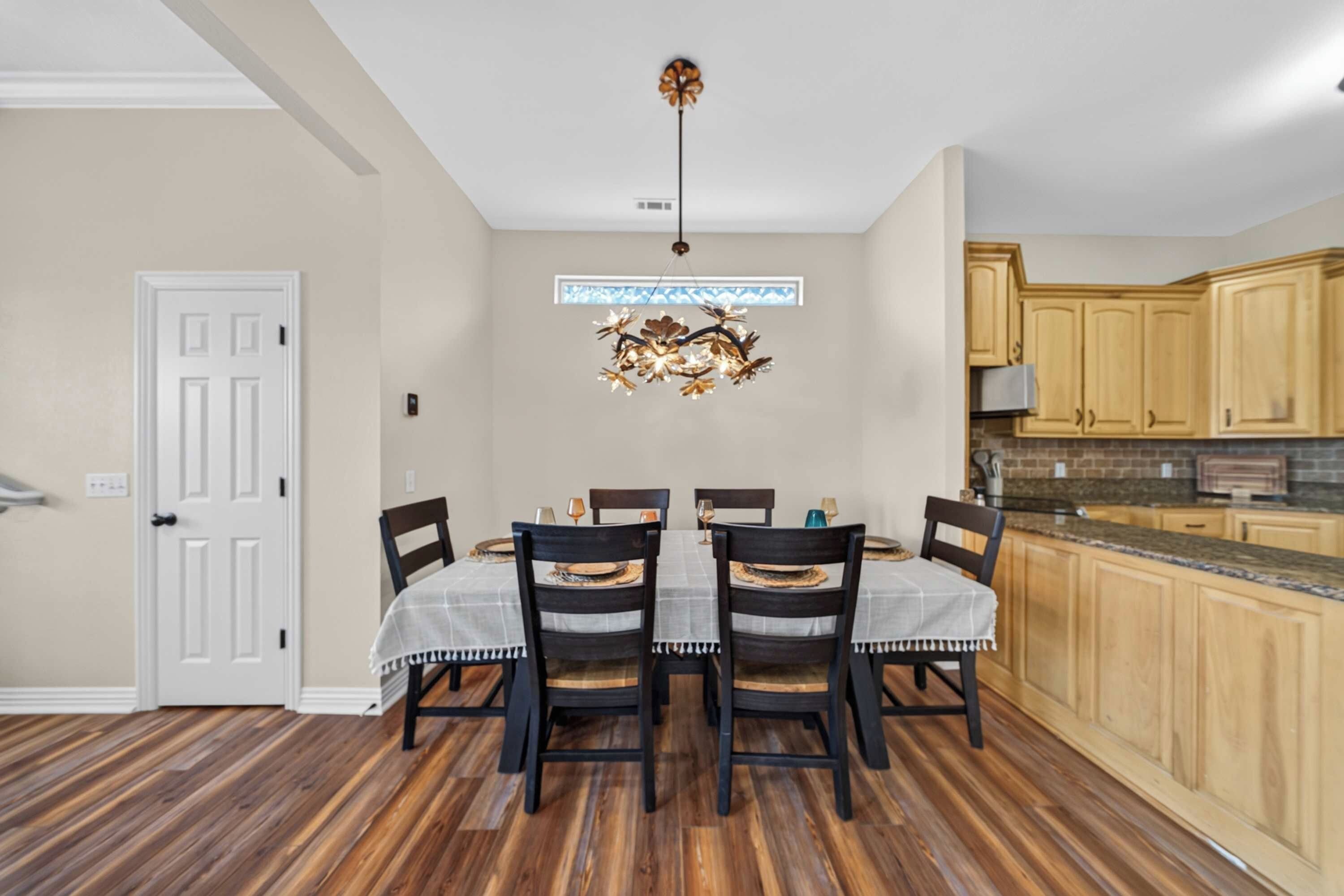 A dining area with a table set for four, wooden chairs, a chandelier, and a kitchen with wooden cabinets.