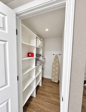 A walk-in closet with white shelving, a red item on the top shelf, and an ironing board against the wall.