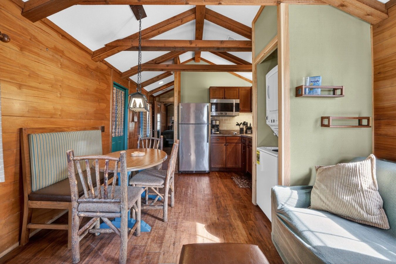 A wooden cabin interior with a dining table, chairs, a sofa, a kitchen area with appliances, and a ceiling with wooden beams.