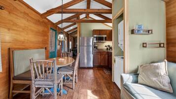 A wooden cabin interior with a dining table, chairs, a sofa, a kitchen area with appliances, and a ceiling with wooden beams.