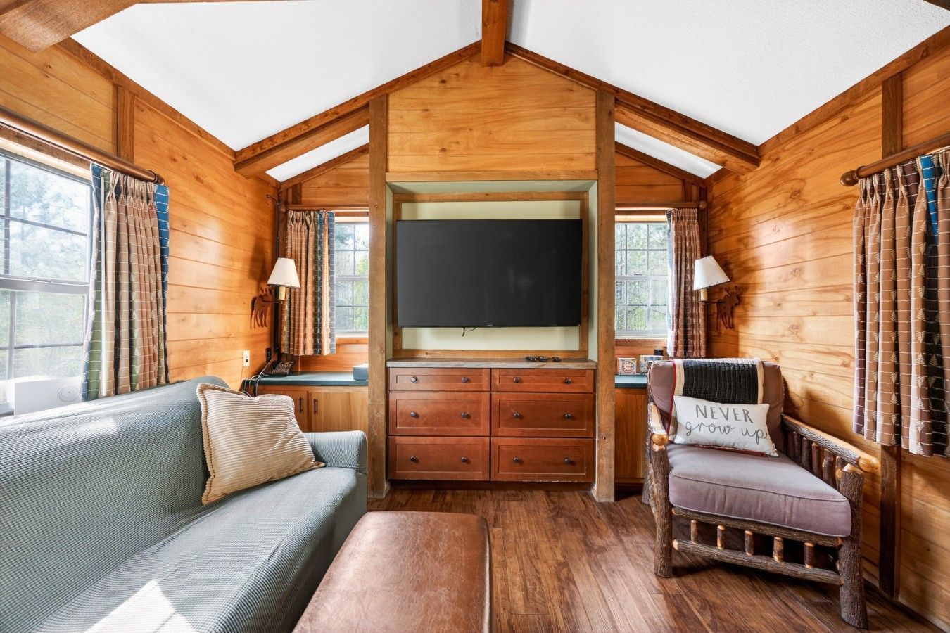 Tunnelmallinen mökin sisustus, jossa on puinen katto, litteänäyttöinen televisio, sohva, nojatuoli ja puinen kaappi.
