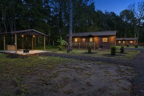 A rustic cabin with a covered patio and a small deck, surrounded by a forested area.