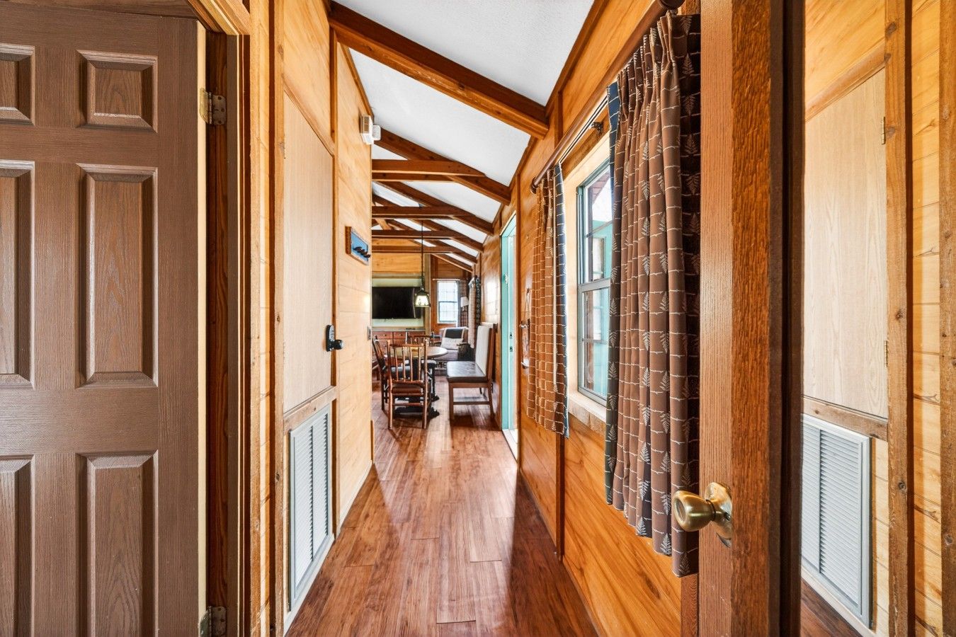 A wooden hallway with a ceiling fan, a dining area with chairs and a table, and a window with curtains.
