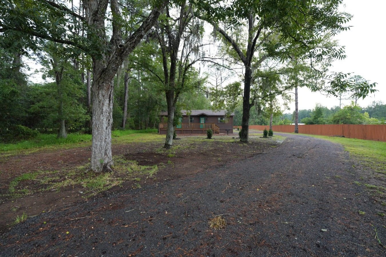 A gravel driveway leading to a wooden cabin with a fenced yard and trees.