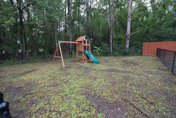 A wooden swing set with a slide in a wooded backyard.
