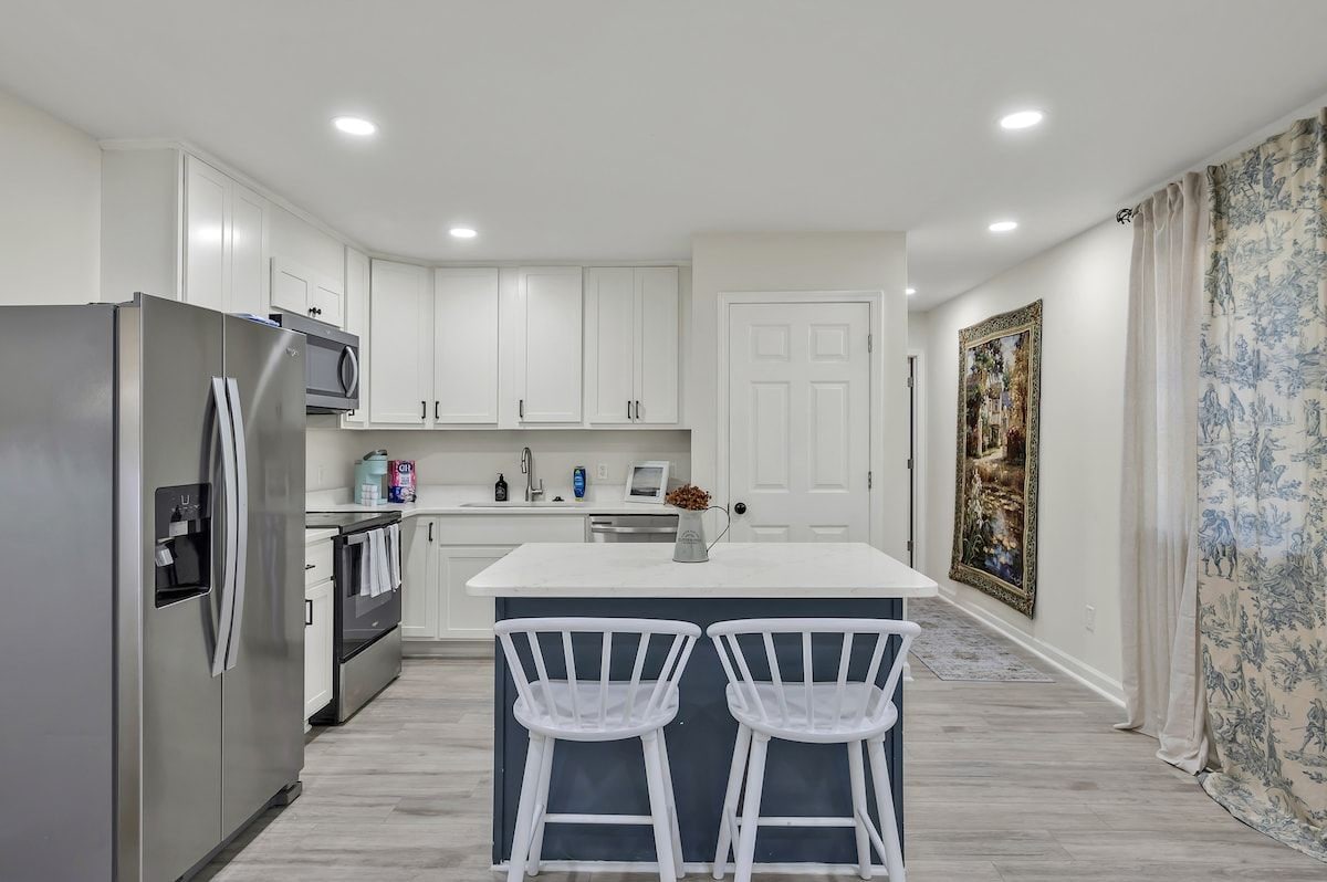 A modern kitchen with white cabinets, a stainless steel refrigerator, a microwave, and a sink. There are two white bar stools at the island.