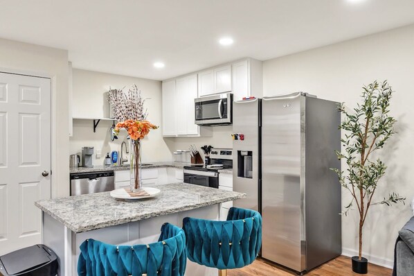 A modern kitchen with white cabinets, a stainless steel refrigerator, a microwave, and a sink. There is a small island with a vase of flowers and two blue chairs. - 8 Min to Duke + DT | Free Parking + Wi-Fi (Durham)