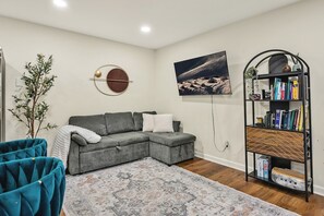 A living room with a grey sectional sofa, a patterned rug, a bookshelf, and a wall-mounted TV. - 8 Min to Duke + DT | Free Parking + Wi-Fi (Durham)