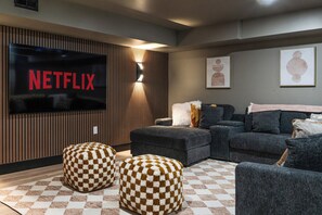 Un soggiorno moderno con una grande TV a schermo piatto che mostra 'NETFLIX' in rosso, un divano componibile, due pouf decorati e un tappeto con motivi.