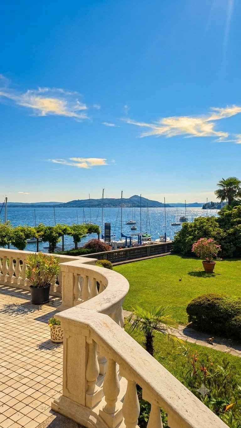 Una terraza empedrada con barandilla, zona ajardinada y vista al mar con barcos.