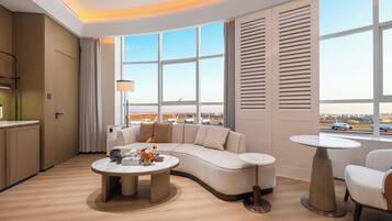 A modern hotel room with a curved sofa, a round coffee table, a small round table, and a view of the airport.