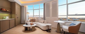 A modern hotel room with a curved sofa, a round coffee table, a small round table, and a view of the airport.