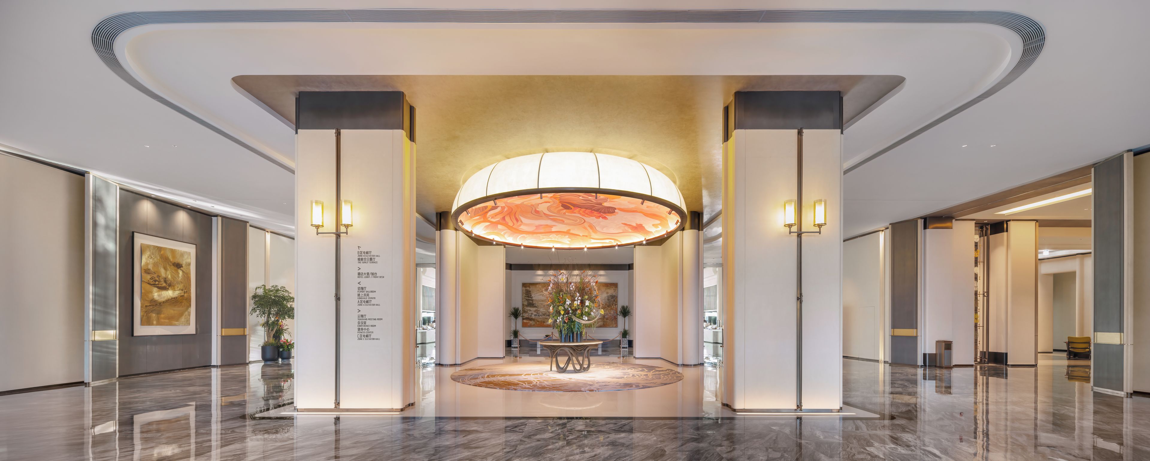 A modern lobby with a central reception desk, a large chandelier, and a polished marble floor.