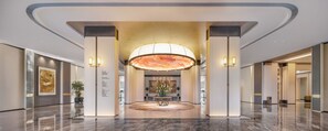 A modern lobby with a central reception desk, a large chandelier, and a polished marble floor.
