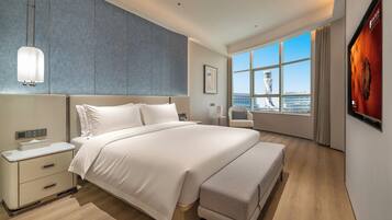 A modern hotel room with a large bed, a bedside table, a chair, and a view of a cityscape through a large window.
