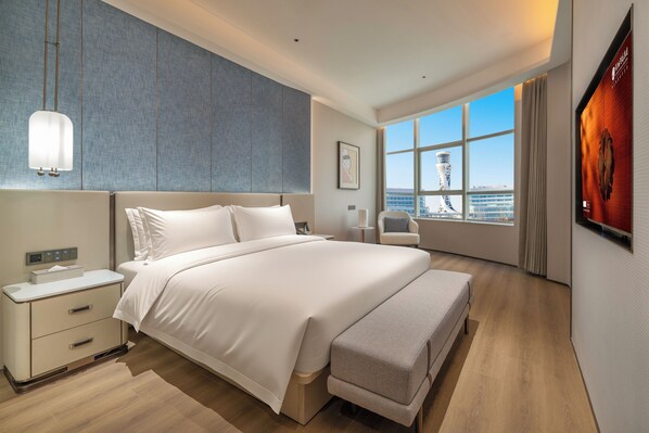 A modern hotel room with a large bed, a bedside table, a chair, and a view of a cityscape through a large window.
