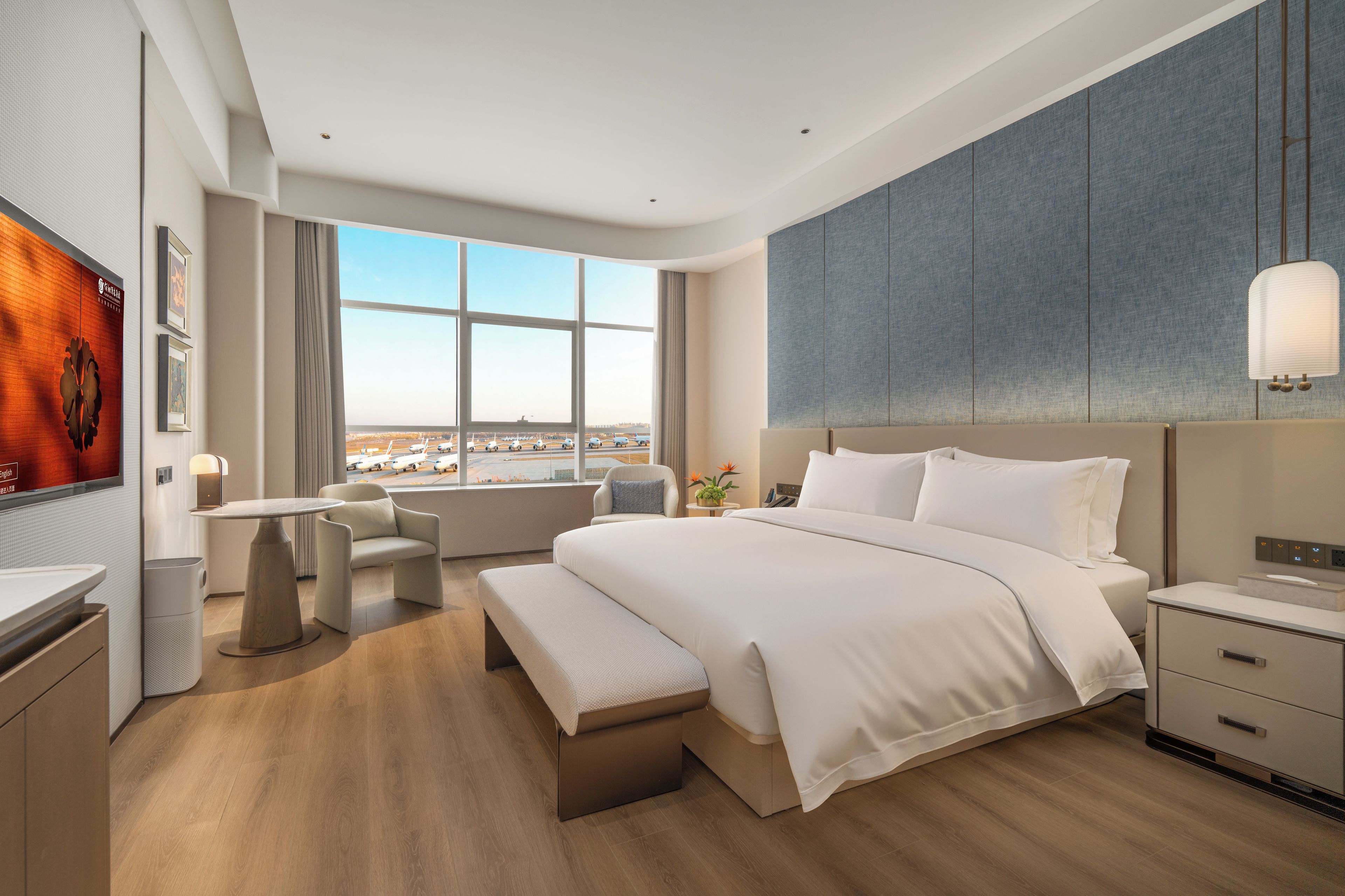 A modern hotel room with a large bed, a desk, and a view of the airport.
